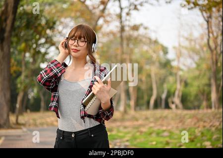 Felice e bella giovane studentessa asiatica in abiti casual e occhiali ama ascoltare la musica attraverso le sue cuffie mentre cammina dentro Foto Stock