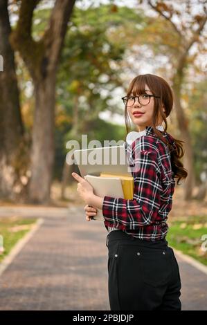 Ritratto di attraente giovane studentessa asiatica in camicia flanella indossando occhiali, tenendo il suo computer portatile e libro, camminando lungo il parco del campus Foto Stock