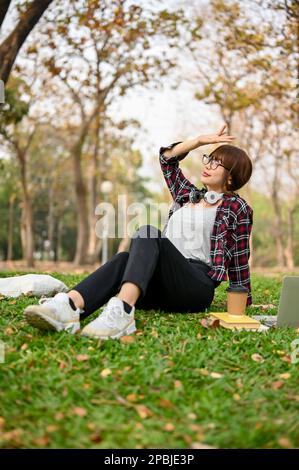 Ritratto di una bella e rilassata giovane donna asiatica in abiti casual, godendo la natura e l'aria fresca mentre si siede sull'erba nel verde par Foto Stock