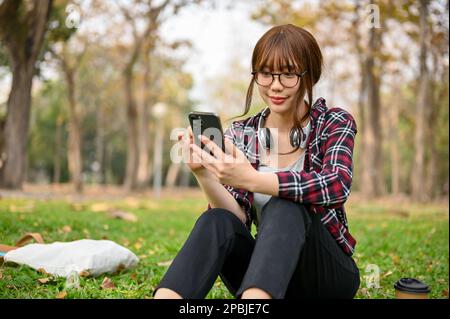 Affascinante e bella giovane studentessa asiatica in abiti casual usa il suo telefono e si rilassa mentre si siede sull'erba in un bellissimo parco verde. Foto Stock