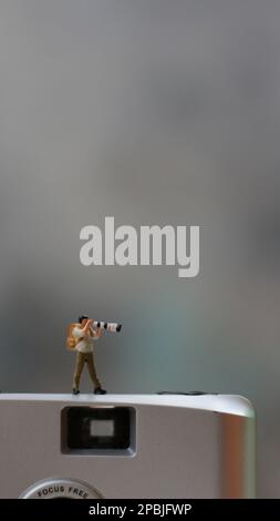 una figura in miniatura che fotografa con una fotocamera contro una fotocamera reale sullo sfondo. Foto Stock