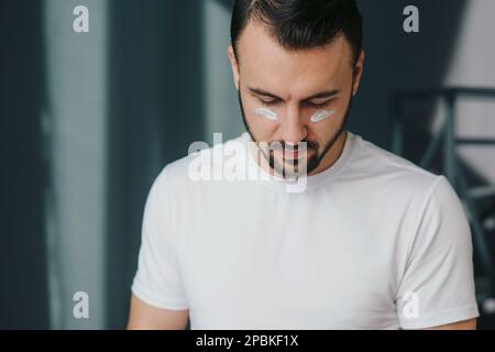 Uomo serio sopportato che applica la maschera di bellezza sul suo viso, avendo trattamento dell'acne dopo il bagno. Problema della pelle, bellezza e concetto di igiene. Foto Stock