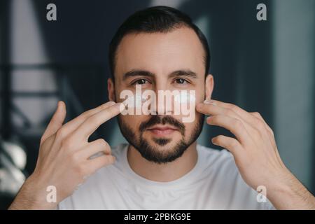 Primo piano ritratto di bellezza di giovane uomo bello in posa in casa accogliente interno e guardando alla fotocamera, mentre applicare crema sulle guance da rughe e. Foto Stock
