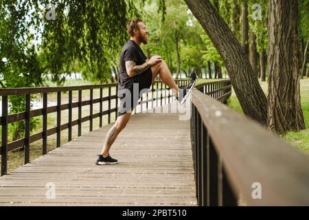 Giovane bell'uomo sorridente tatuato bearded allungando la gamba su ringhiera di legno parco soleggiato Foto Stock