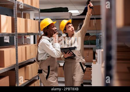 Due giovani addetti al magazzino che utilizzano apparecchiature per lo scanner di codici a barre per la supervisione delle scorte. Donne afro-americane e caucasiche che indossano caschi protettivi scansione scatole di cartone Foto Stock