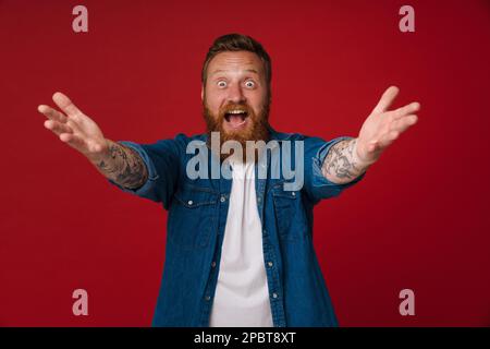 Adulto bearded tatuato bell'entusiasta redhead uomo con le braccia aperte guardando la macchina fotografica, mentre in piedi sopra isolato sfondo rosso Foto Stock