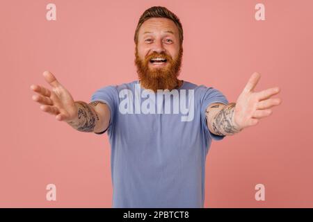 Adulto arrossato tatuato sorridente uomo felice con braccia aperte guardando la macchina fotografica, mentre in piedi su isolato sfondo rosa Foto Stock