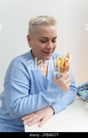 Ritratto di donna bianca adulta con capelli corti tenendo in mano la pita greca souvlaki. Allegra ragazza Ucraina gustando il pranzo in fast food ristorante Foto Stock