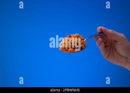 Bulgur cotto con pomodori a dadini su un cucchiaio d'argento tenuto da mano maschio caucasica. Primo piano ripresa in studio, isolata su sfondo blu. Foto Stock