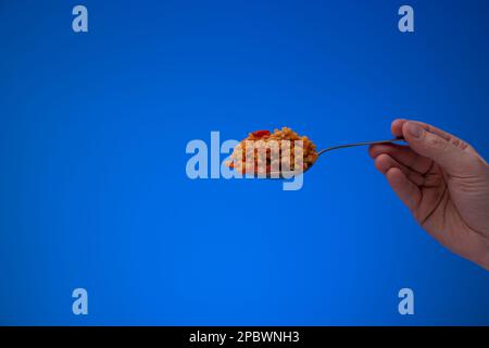 Bulgur cotto con pomodori a dadini su un cucchiaio d'argento tenuto da mano maschio caucasica. Primo piano ripresa in studio, isolata su sfondo blu. Foto Stock