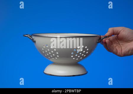 Colander da cucina vuoto rotondo in metallo bianco tenuto da mano maschile. Primo piano ripresa in studio, isolata su sfondo blu. Foto Stock