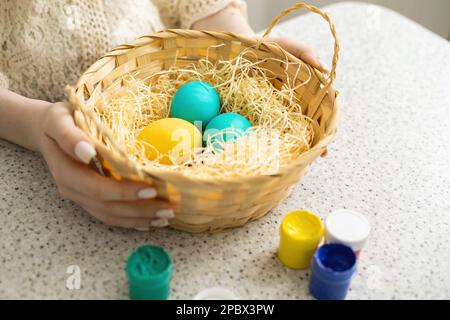 Un primo piano delle mani di una ragazza che tiene un cesto di vimini contenente uova dipinte. Ci sono pitture sul tavolo vicino. Pasqua Foto Stock