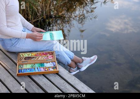 Donna disegno con morbidi pastelli su molo di legno vicino al fiume, primo piano Foto Stock