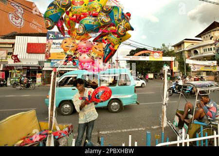 Un fornitore di palloncini mobili e un autista di risciò in un contesto di traffico stradale su Penghibur Street, una strada costiera con attività economiche molto trafficate a Makassar City, Sulawesi meridionale, Indonesia. Una delle 30 città costiere dell'Indonesia che si stima sia potenzialmente colpita dall'aumento del livello del mare, Makassar è stata considerata dai ricercatori e dagli scienziati una città con notevoli problemi ambientali e sociali; principalmente come risultato della condizione geomorfologica e della politica di utilizzazione del territorio e di sviluppo costiero, in particolare i progetti di bonifica attualmente in corso. Foto Stock