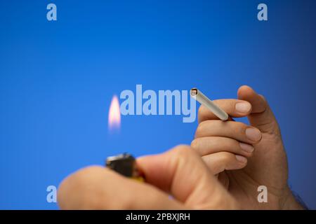 Una sigaretta e un accendisigari spenti tenuti in mano da una mano maschio caucasica. Primo piano ripresa in studio, isolata su sfondo blu. Foto Stock