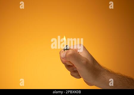 Accendino e fiamma accesi tenuti in mano da una mano maschio caucasica. Primo piano ripresa in studio, isolata su sfondo arancione. Foto Stock