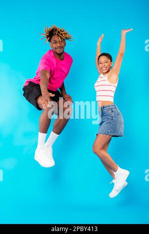 Tutta la giovane coppia multirazziale gioiosa in abiti eleganti sorridendo brillante mentre si salta su sfondo blu e si guarda la macchina fotografica Foto Stock