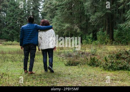 Vista posteriore corpo pieno di irriconoscibile madre e figlio in abiti caldi camminando in legno verde con alberi di conifere e abbraccio Foto Stock