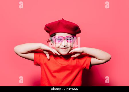 Ragazza giovane divertente in abito casual rosso alla moda tenendo le mani vicino al viso guardando la macchina fotografica e in piedi su sfondo rosa Foto Stock