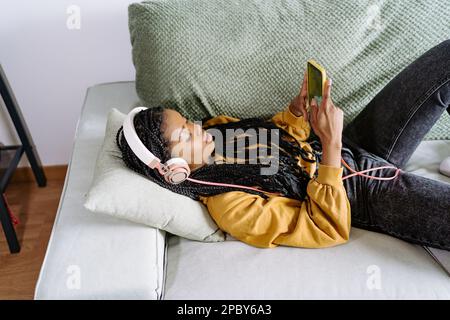Dall'alto della giovane afro-americana amena non emotiva in abiti casual con trecce sdraiate su un comodo divano e navigare in internet su smartphone whi Foto Stock