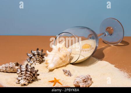 Dall'alto di wineglass posto su un tavolo beige con sabbia rovesciato e varie conchiglie vicino alle stelle marine su sfondo blu Foto Stock