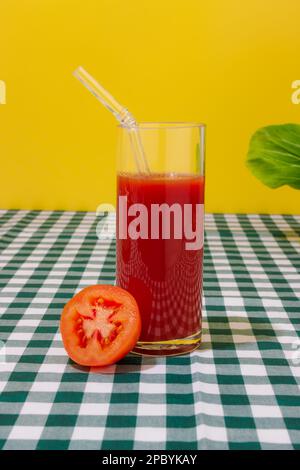 Bicchiere trasparente di succo di pomodoro fresco con cannuccia di vetro posta su tovaglia a scacchi su fondo giallo Foto Stock