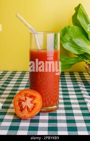 Bicchiere trasparente di succo di pomodoro fresco con cannuccia di vetro posta su tovaglia a scacchi su fondo giallo Foto Stock