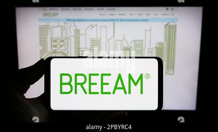 Persona che tiene lo smartphone con il logo della certificazione verde edificio BREEAM sullo schermo di fronte al sito Web. Messa a fuoco sul display del telefono. Foto Stock