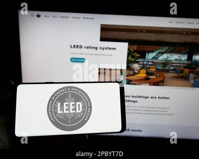 Persona che tiene in mano uno smartphone con il logo della certificazione US Green Building LEED sullo schermo di fronte al sito Web. Messa a fuoco sul display del telefono. Foto Stock