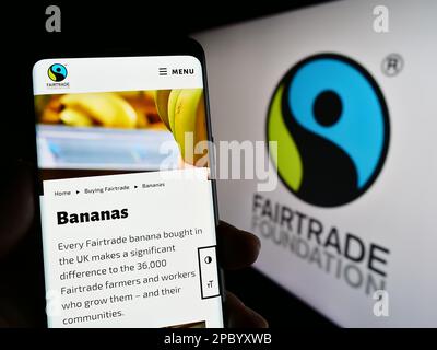 Persona che tiene uno smartphone con il sito web dell'organizzazione benefica la Fairtrade Foundation sullo schermo con il logo. Messa a fuoco al centro del display del telefono. Foto Stock