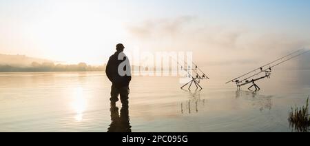Avventure di pesca, pesca alla carpa. Pescatore con stivali sul lago al sorgere del sole in una mattina misty è la pesca con la tecnica di pesca al carpfishing Foto Stock