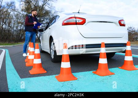Istruttore vicino all'auto con studente durante l'esame alla pista di prova della scuola guida, focalizzato sui coni stradali Foto Stock
