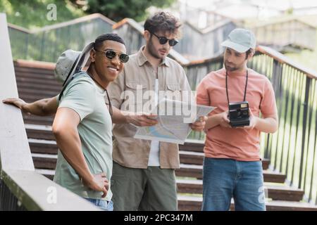 viaggiatore afro-americano spensierato sorridente alla macchina fotografica mentre si trova vicino agli amici interracial con la mappa sulle scale della strada della città, immagine di scorta Foto Stock