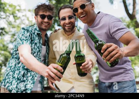 uomini multietnici spensierati ed eleganti in occhiali da sole che si aggraffano le bottiglie di birra nel parco, immagine stock Foto Stock