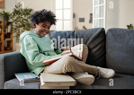 Buon scolaro in felpa con cappuccio e pantaloni facendo appunti in copybook mentre si siede sul divano e preparando l'attività a casa per la lezione Foto Stock