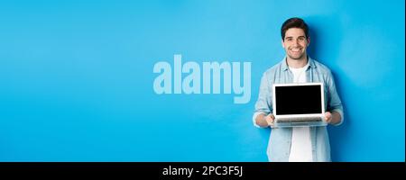 Bello giovane uomo introdurre il prodotto sullo schermo del laptop, mostrando il computer e sorridendo, in piedi su sfondo blu Foto Stock