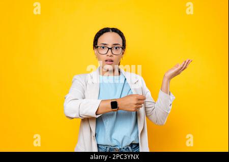 Perplesso confusa donna d'affari brasiliana, dipendente aziendale, utilizzando l'orologio intelligente sulla sua mano, preoccupato per essere in ritardo, si trova su sfondo arancione isolato, guarda la fotocamera dispiaciuta, gesturando la mano Foto Stock