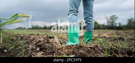 Primo piano stivali di contadino che lavora sul campo. Turbine eoliche all'orizzonte Foto Stock