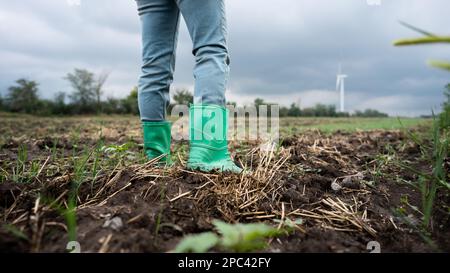 Primo piano stivali di contadino che lavora sul campo. Turbine eoliche all'orizzonte Foto Stock