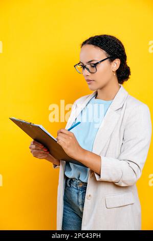 Foto verticale di una donna ispanica focalizzata di successo, in abiti alla moda, hr manager, agente immobiliare, tiene e firma documenti, concentrato li guarda, stand su sfondo giallo isolato Foto Stock