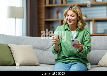 Sorridente donna anziana bella seduta sul divano a casa. Tenere in mano un telefono, utilizzando una carta di credito. Sorridendo effettua acquisti online, apre un account, assegni. Foto Stock