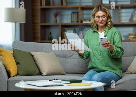 Lavoro da casa. Una donna anziana in occhiali è seduta sul divano a casa, tenendo un telefono e documenti in mano. Funziona con documenti, assegni e pagamenti online fatture. Foto Stock