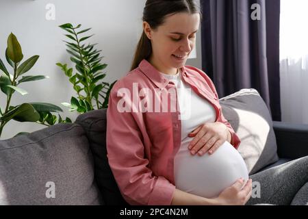 Buon concetto di maternità. Sorridente giovane donna incinta seduta su un comodo divano in camera leggera toccare grande pancia godere le fasi tardive della gravidanza. Carin Foto Stock