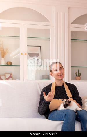 Giovane uomo positivo in abiti casual con braccio rotto in sling parlando mentre seduto sul divano con gatto nel soggiorno a casa Foto Stock