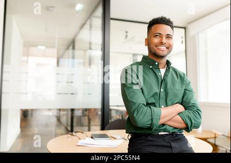 L'ambizioso imprenditore indiano indiano ad alto livello si trova in uno spazio ufficio contemporaneo con le braccia incrociate appoggiate sulla scrivania dell'ufficio, l'uomo d'affari latino in camicia casual guarda con il sorriso Foto Stock
