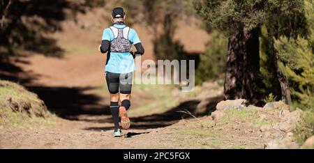 Atleta che corre la maratona di montagna lungo un sentiero forestale con uno zaino e bastoni da passeggio Foto Stock