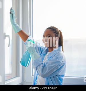 Bella giovane donna nera sorridente mentre pulisce la finestra a casa Foto Stock
