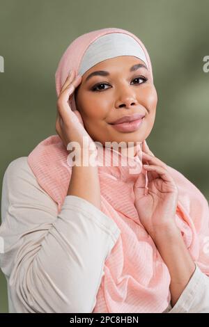 donna musulmana multirazziale positiva in hijab rosa che posa con le mani vicino al viso su sfondo verde, immagine stock Foto Stock