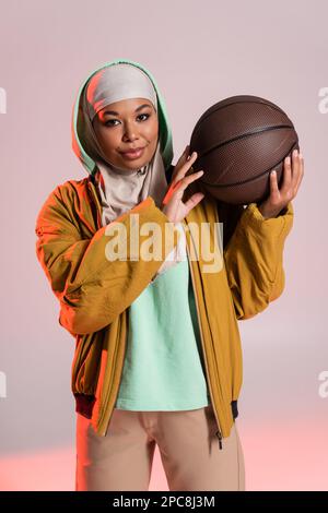 bella donna multirazziale in giacca gialla e hijab tenendo il basket mentre sorridendo alla macchina fotografica su sfondo grigio e rosa, immagine stock Foto Stock