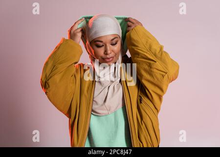 giovane e trendy donna multirazziale in hijab e gialle bomber giacca con cappuccio su sfondo grigio rosa, immagine stock Foto Stock